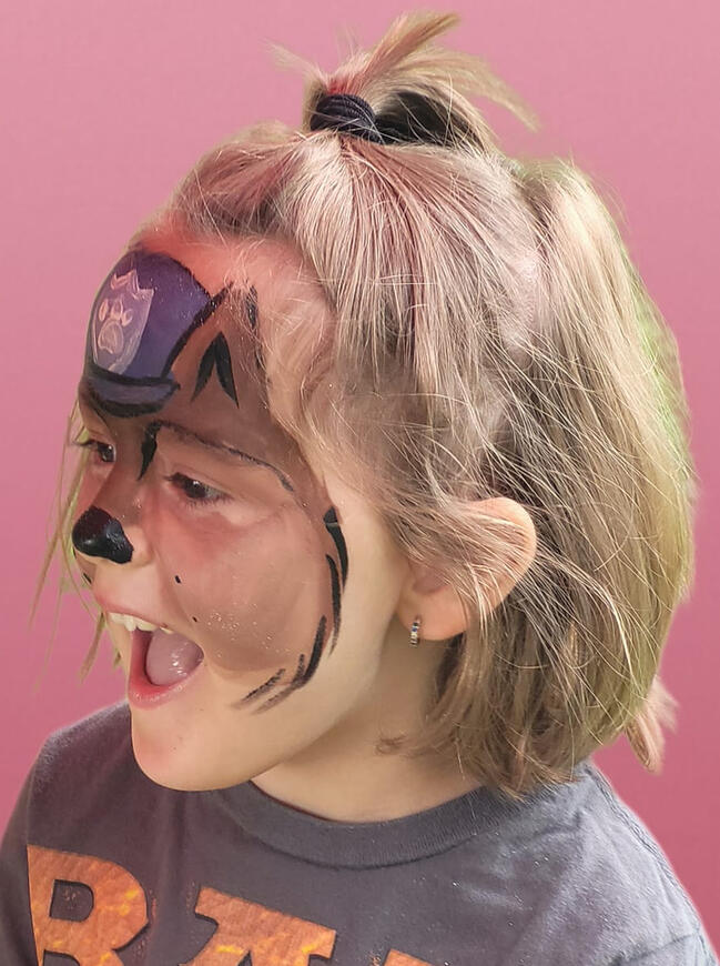 young girl smiling, looking at her reflection. her face is painted like a police dog