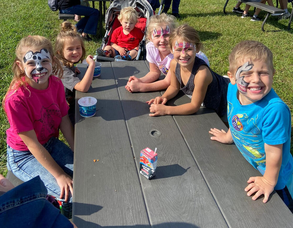 group of children at a picnic table, smiling, several have their faces painted